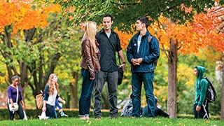 A photo of three students talking on campus with other students in the background.