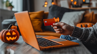 Woman holding a credit card in front of an open laptop beside a jack-o’-lantern.
