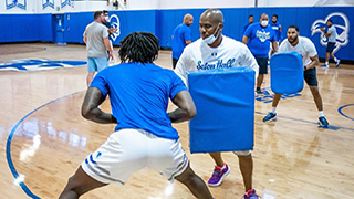 Seton Hall Pirates men's basketball head coach Shaheen Holloway smiling while coaching.Shaheen Holloway practicing with Kadary Richmond of the Pirates basketball team in the gym while holding a pad.