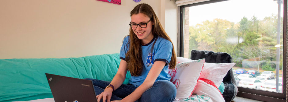 Student sitting on a bed in a dorm room looking at a computer. 